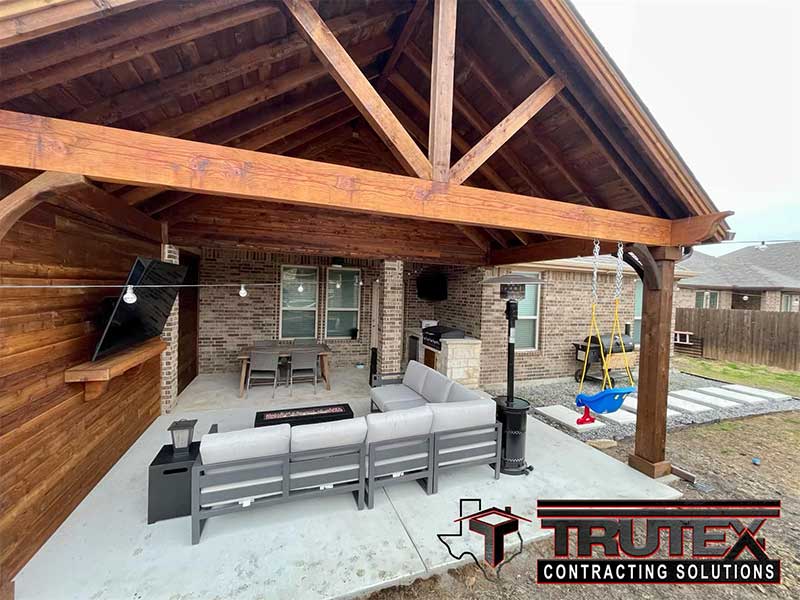 Outdoor patio area with a roof that has sofas, chairs and table, a bar-b-q, a small swing set, and a large television